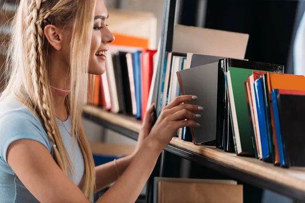 Усміхнена молода жінка вибирає книгу на книжковій полиці в бібліотеці — стокове фото