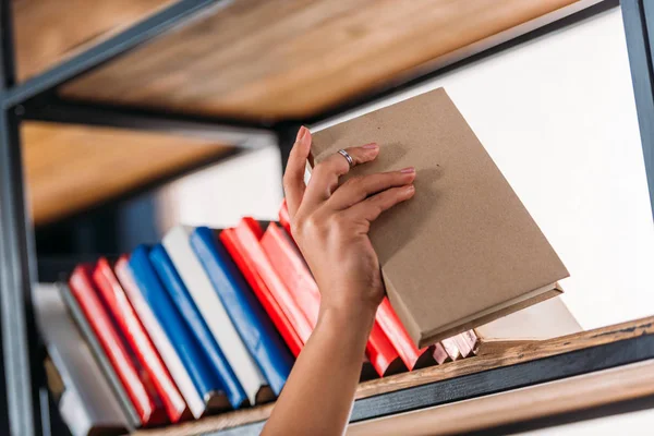 Recorte de estudiante sosteniendo libro en estantería en la biblioteca - foto de stock
