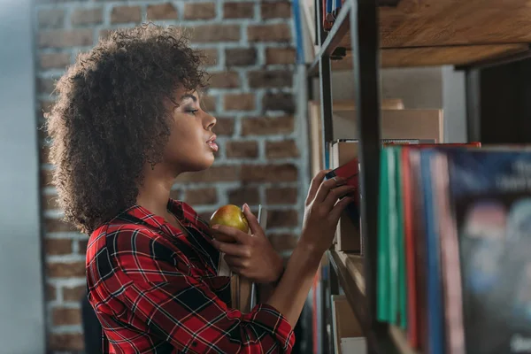 Side view of beautiful african american student holding books with apple and looking at bookshelves — Stock Photo