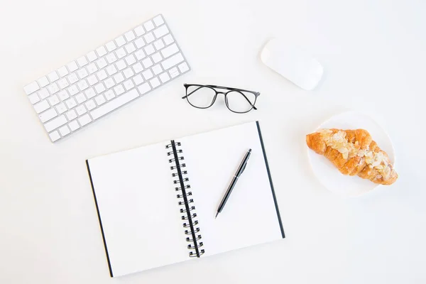 Draufsicht auf leckeres Croissant auf Teller, leeres Notizbuch mit Stift, Tastatur und Computermaus am Arbeitsplatz — Stockfoto