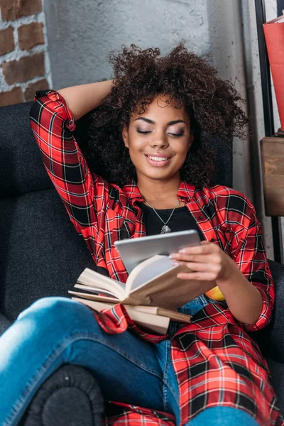 Усміхнена молода жінка сидить у кріслі з книгами і використовує цифровий планшет — стокове фото
