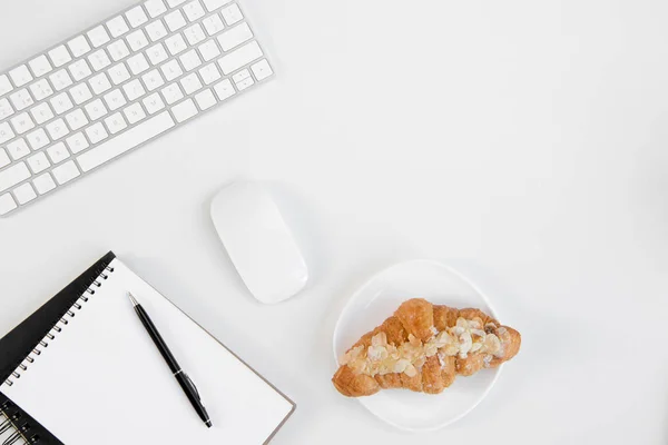 Vista dall'alto del gustoso croissant sul piatto, quaderno bianco con penna, tastiera e mouse sul posto di lavoro — Foto stock