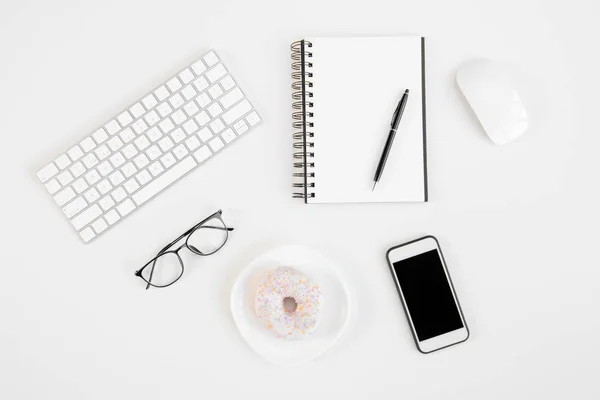 Vista superior del teléfono inteligente con pantalla en blanco, cuaderno en blanco con pluma, anteojos, teclado, ratón de la computadora y rosquilla sabrosa en el lugar de trabajo - foto de stock