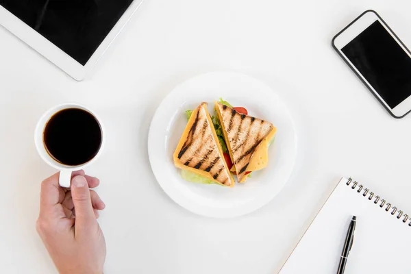 Top view of person eating sandwiches and drinking coffee at workplace with electronics — Stock Photo