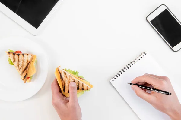 Ansicht von oben: Person schreibt in Notizbuch, während sie am Arbeitsplatz Sandwiches isst — Stockfoto