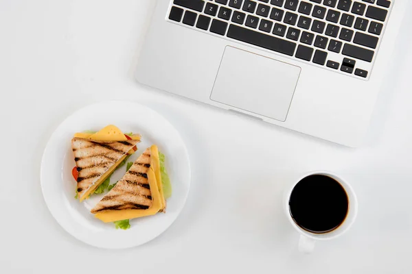 Vista superior de sabrosos sándwiches en el plato, taza de café y computadora portátil en el lugar de trabajo — Stock Photo