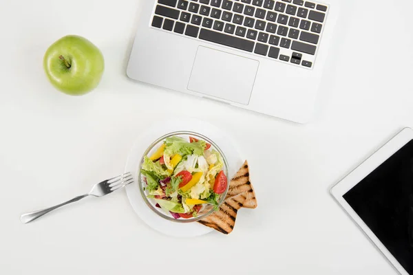 Vista superior del portátil y la tableta digital con pantalla en blanco, manzana verde fresca y ensalada fresca con tostadas en el lugar de trabajo - foto de stock