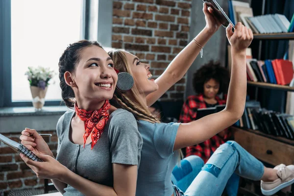 Smiling young women sitting back to back while writing in notebook and using digital tablet — Stock Photo