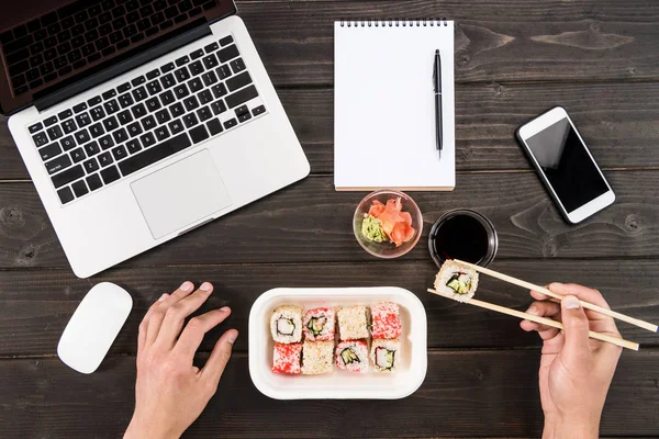 Vue de dessus des mains humaines mangeant des sushis pendant le travail avec des appareils numériques sur une table en bois — Photo de stock