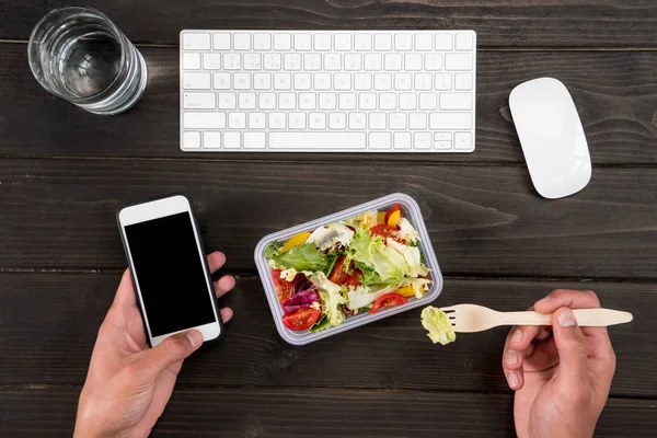 Top view of human hand eating fresh salad and using smartphone during work. — Stock Photo