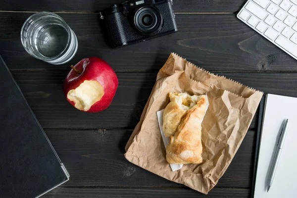 Top view of bread loaf with apple and camera with notebook and glass of water on tabletop — Stock Photo