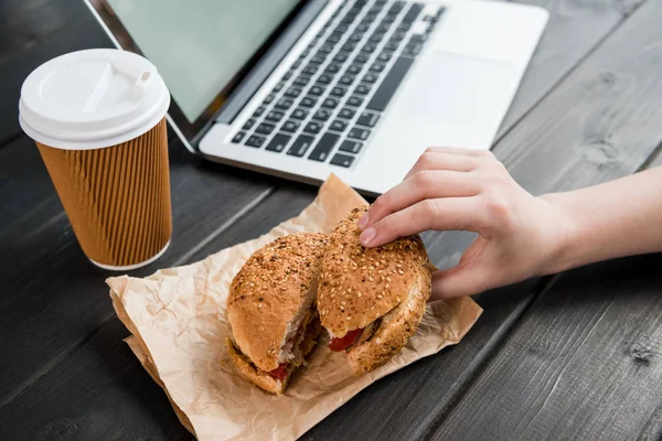 Gros plan de main tenant hamburger avec café pour aller et ordinateur portable sur table en bois — Photo de stock
