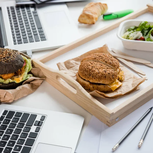 Primer plano de sabrosas hamburguesas con ensalada fresca y pan de pan con computadoras portátiles en la mesa - foto de stock