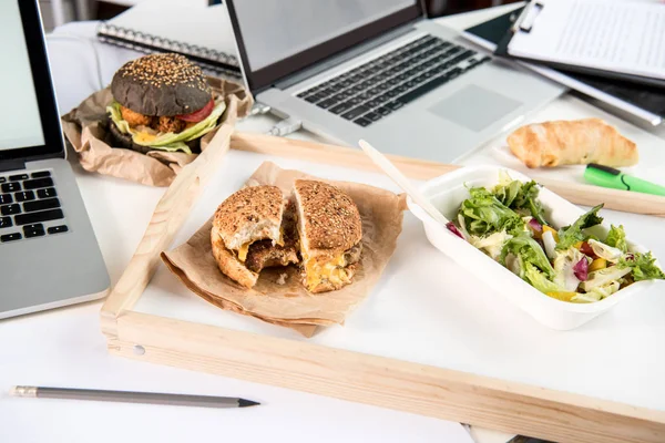 Hambúrgueres saborosos com salada fresca e pão com laptops na mesa — Fotografia de Stock