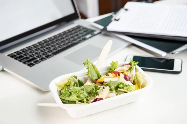 Primer plano de ensalada fresca con computadora portátil y teléfono inteligente en la mesa en el lugar de trabajo - foto de stock