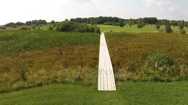 Беспилотник пролетает над зеленым полем для гольфа с деревянным мостом через болото — стоковое видео