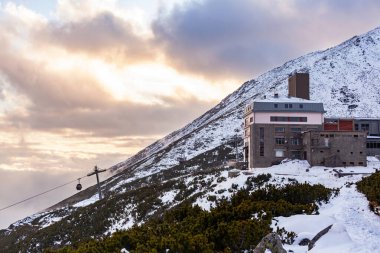 Bulutların üstündeki dağın tepesinde bir taş bina duruyor.