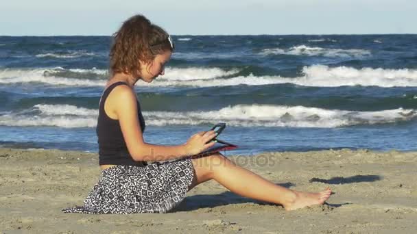 Piękna młoda kobieta siedzi na plaży oraz składowanie na jej komputerze tablet pc w letni dzień — Wideo stockowe