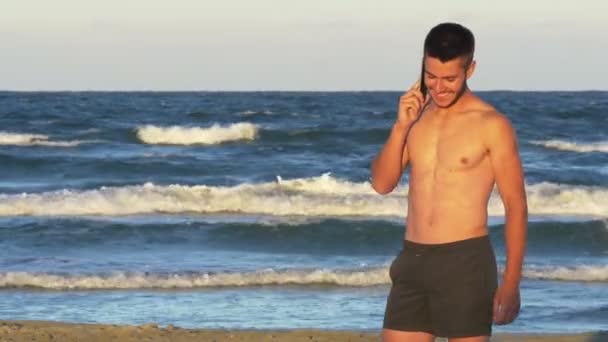 Pohledný mladý muž mluví po telefonu s mořem jako pozadí — Stock video