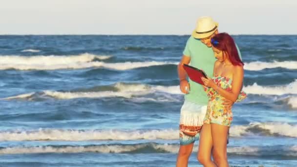 Νεαρό ζευγάρι έχει τη διασκέδαση σε μια καλοκαιρινή μέρα δίπλα στη θάλασσα με ένα tablet — Αρχείο Βίντεο