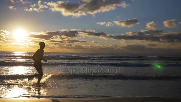 Homem correndo na costa em uma praia de areia salpicando com água em câmera lenta — Vídeo de Stock