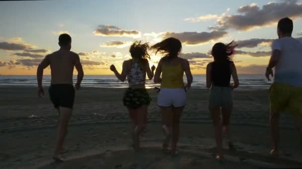 Grupo de jóvenes amigos corriendo al agua desde la playa al atardecer — Vídeos de Stock