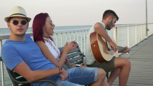 Tři přátelé sedí na lavičce s mořem jako pozadí a zpívání s kytarou — Stock video