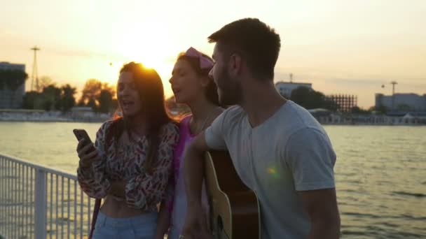 Drie vrienden zingen met een gitaar op een brug over de zee op zomertijd — Stockvideo