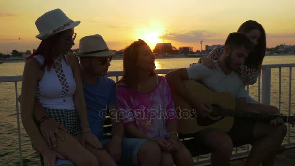 Grupo de amigos sentados en un banco y cantando con una guitarra al atardecer en un pontón — Vídeos de Stock