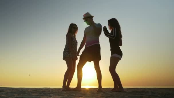 Młody człowiek tańczy na plaży z dwoma kobietami o wschodzie słońca — Wideo stockowe