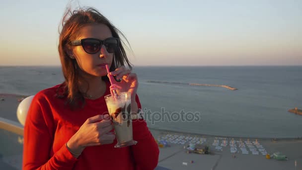 Junge Teenagerin trinkt eine Frappe auf einer Terrasse mit Meerblick — Stockvideo