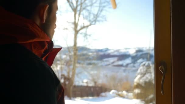 Νεαρός άνδρας βλέπει έξω από το παράθυρο στο όμορφο τοπίο το χειμώνα τα βουνά — Αρχείο Βίντεο