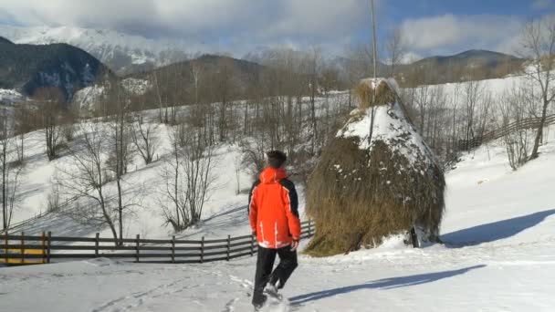 Чоловік виходить на вулицю на гору в сільській місцевості через сніг вздовж стопи сіна — стокове відео