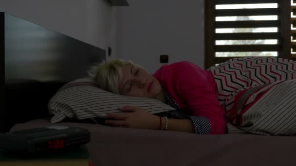 Молодая женщина спит в постели утром и поворачивается, глядя на будильник — стоковое видео