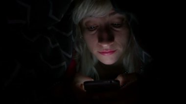 Yatakta battaniyenin altında akıllı telefon kullanarak ve gülümseyen neşeli genç kadın