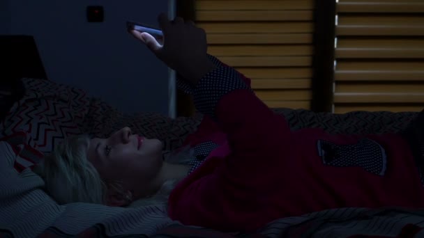 Mujer de pie en su cama por la noche riendo mientras navega por Internet en su teléfono inteligente — Vídeo de stock