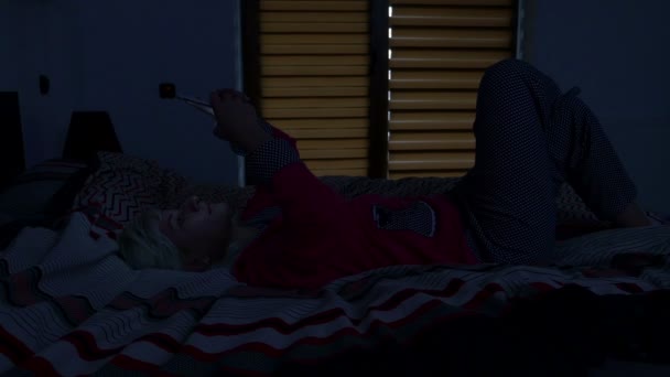 彼女のスマート フォンを見て、笑いながら夕方にはベッドで横になっている女性 — ストック動画