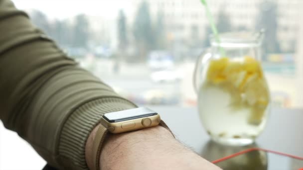 Nahaufnahme eines Mannes, der auf der Smartwatch nach Benachrichtigungen sucht, während er mit Limonade am Tisch sitzt — Stockvideo