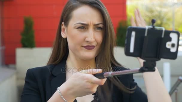 Vista de perto de poses de menina agradável e faz caras engraçadas com câmera de smartphone e vara de selfie na cidade — Vídeo de Stock