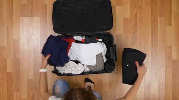 Bovenaanzicht van jong meisje zittend op een houten vloer en uitpakken van kleren uit trolley tas — Stockvideo