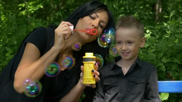 Mãe entretendo e ensinando seu filho fazendo bolhas de sabão iridescentes em um banco no parque — Vídeo de Stock