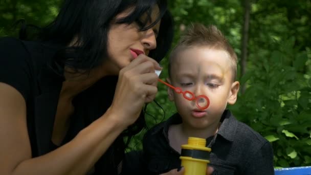 Piccolo figlio che soffia bolle di sapone alla luce del sole con la sua giovane madre nello spazio ricreativo pubblico — Video Stock