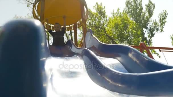 Felice gioioso bambino scivolare giù uno scivolo blu su un parco giochi in un parco — Video Stock