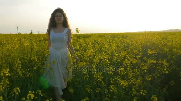 Linda chica con vestido blanco caminando hacia la cámara en el campo de colza flores amarillas — Vídeos de Stock