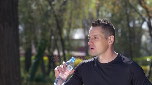 Κοντινό πλάνο του νεαρού αθλητή πόσιμου νερού και λαμβάνοντας το χρόνο μακριά από την κατάρτιση τρέξιμο στο πάρκο σε αργή κίνηση — Αρχείο Βίντεο