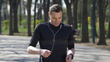 Müzik ve kulaklıklar giyen ve durdurma parkta smartwatch İzleyicisi'ni kontrol etmek için eğitim genç Maratoncu