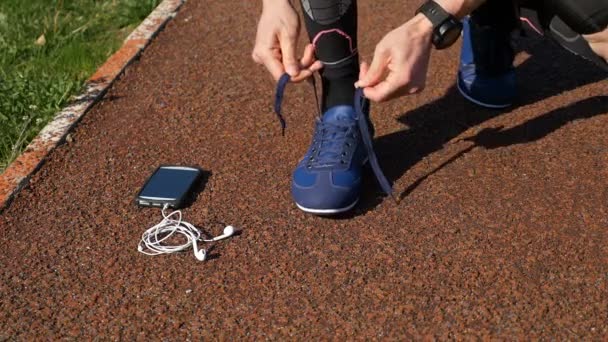 Крупный план бегуна, завязывающего шнурки, затем поднимающего смартфон с помощью бесплатных наушников и бегущего по трассе — стоковое видео