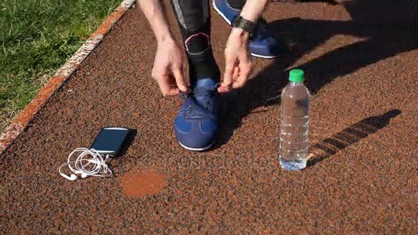 Jogger with smartwatch tying shoelaces and picking up smartphone with earphones and water bottle — Stock Video