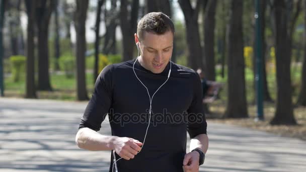 Joven maratonista escuchando música y entrenando en el parque usando auriculares y deteniéndose para revisar el rastreador de relojes inteligentes — Vídeo de stock