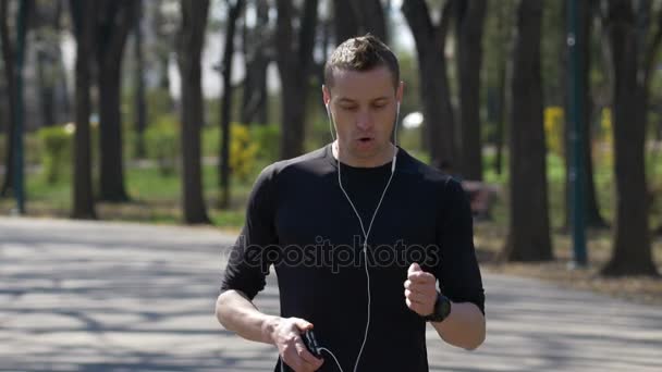 在公园听音乐停下来检查智能手机和 smartwatch 追踪器中运行的男性慢跑 — 图库视频影像